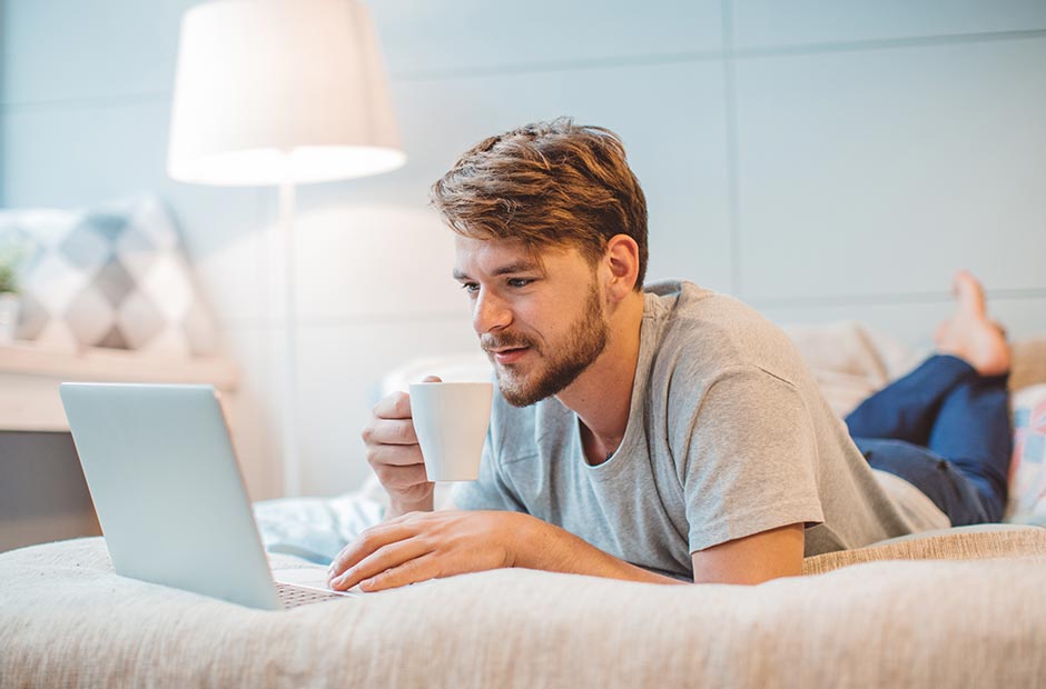 Homme allongé sur son lit buvant une boisson chaude tout en lisant des e-mails sur un ordinateur portable